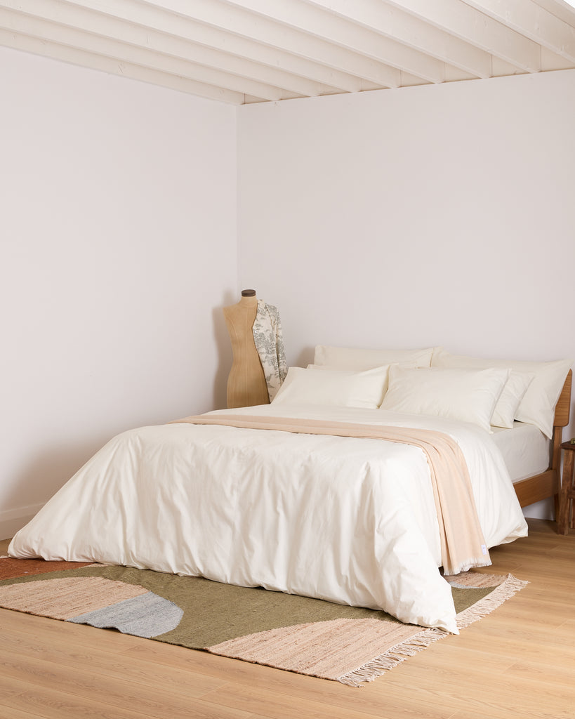 Bedroom with a bed covered in cream bedding and beige blanket, wooden floor, and white walls.